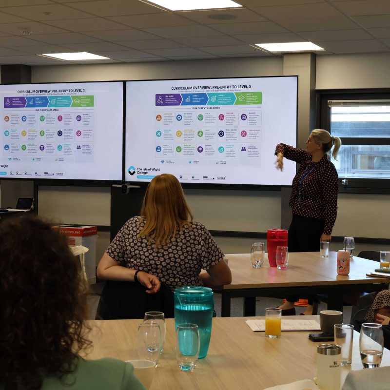 A woman giving a speech to a room gestures towards her powerpoint