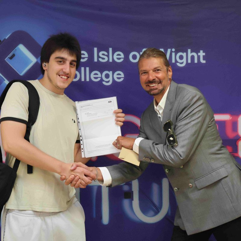 a lecturer shakes the hand of a student who is holding their GCSE results as they pose for a photo.