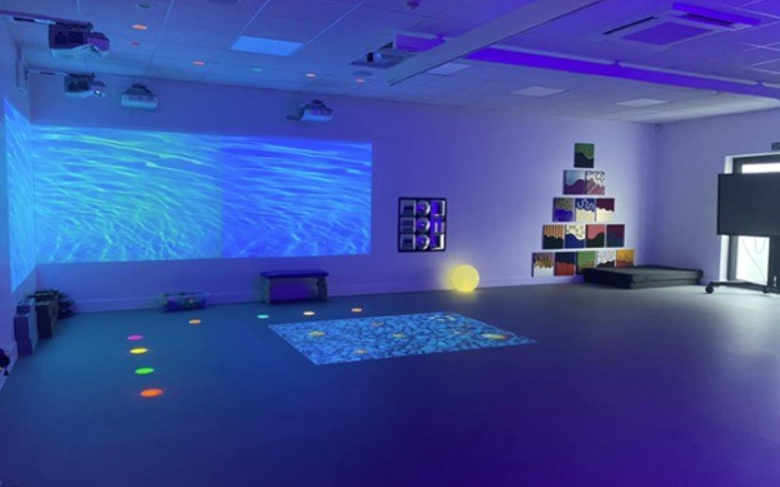 Blue-lit sensory room with interactive water projections, coloured floor lights, sensory wall panels, and soft seating